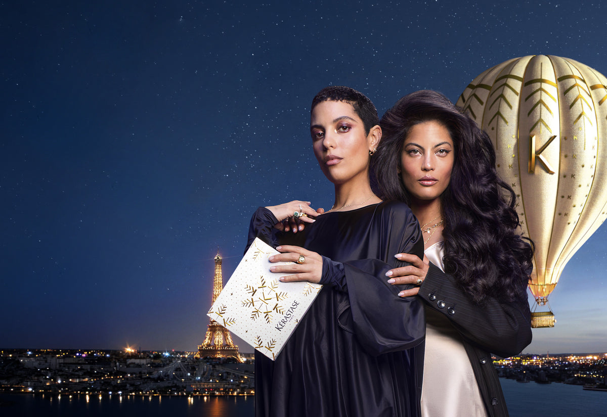 Two women posing together in front of a starry night sky with the Eiffel Tower and a hot air balloon in the background. One woman holds a white box with gold and black lettering that says "Kérastase"
