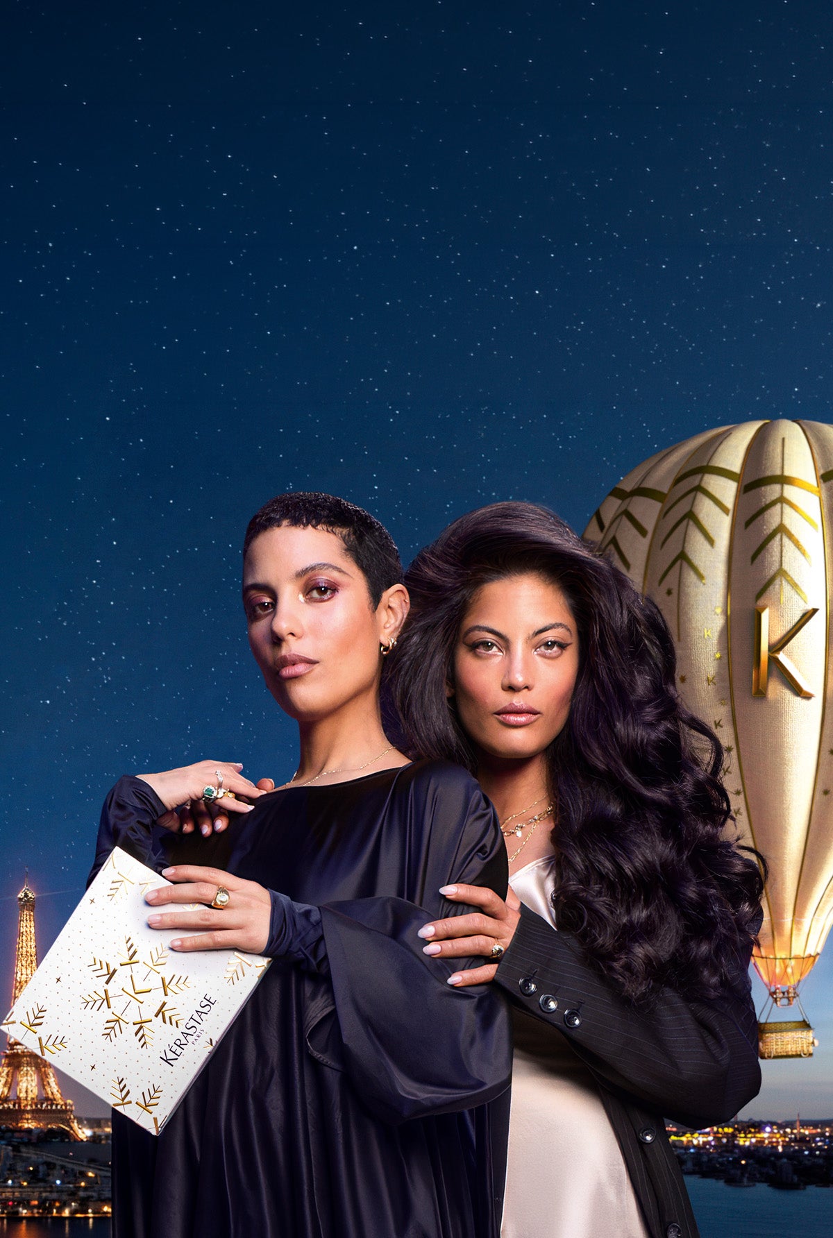 Two women posing together in front of a starry night sky backdrop with a hot air balloon and city lights. One woman wears a black long-sleeve dress and holds a white envelope decorated with gold stars and the word 'AMERICA'.